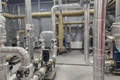 Industrial piping system featuring various insulated pipes, valves, and control panels arranged in mechanical room, showcasing clean and organized layout with metallic surfaces and functional design