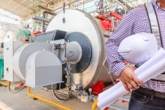 Young engineer holding helmet safety and blueprints working with gas boiler of heating system equipment in a boiler room
