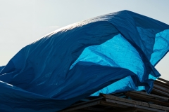 A large blue tarp billows dramatically in the wind, covering construction materials or a roof structure against a bright, sunny sky.