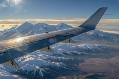 Snow-capped peaks meet a clear blue sky as sunlight dances on the aircraft wing, thin clouds blur the horizon.