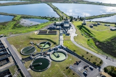 Aerial, wide view of an industrial waste water treatment plant containing several clarifier tanks, stabilization ponds and maturation ponds beyond.