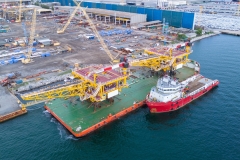 offshore oil and gas platform structure loaded on a heavy transport barge with a supply vessel at a coastal shipyard. Global energy industry and marine construction.