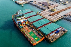 barge heavily loaded with massive steel pipes moored alongside an offshore supply vessel at an industrial port, representing heavy marine construction and project cargo logistics.
