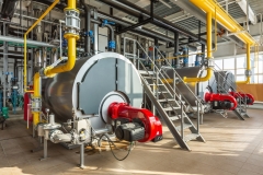 The interior of an industrial boiler room with three large boilers, many pipes, valves and sensors